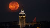 A red full moon is photographed shining next to an illuminated medieval tower at night.