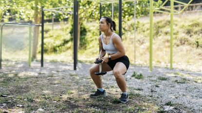 Woman doing squat while holding a dumbbell outside in a park