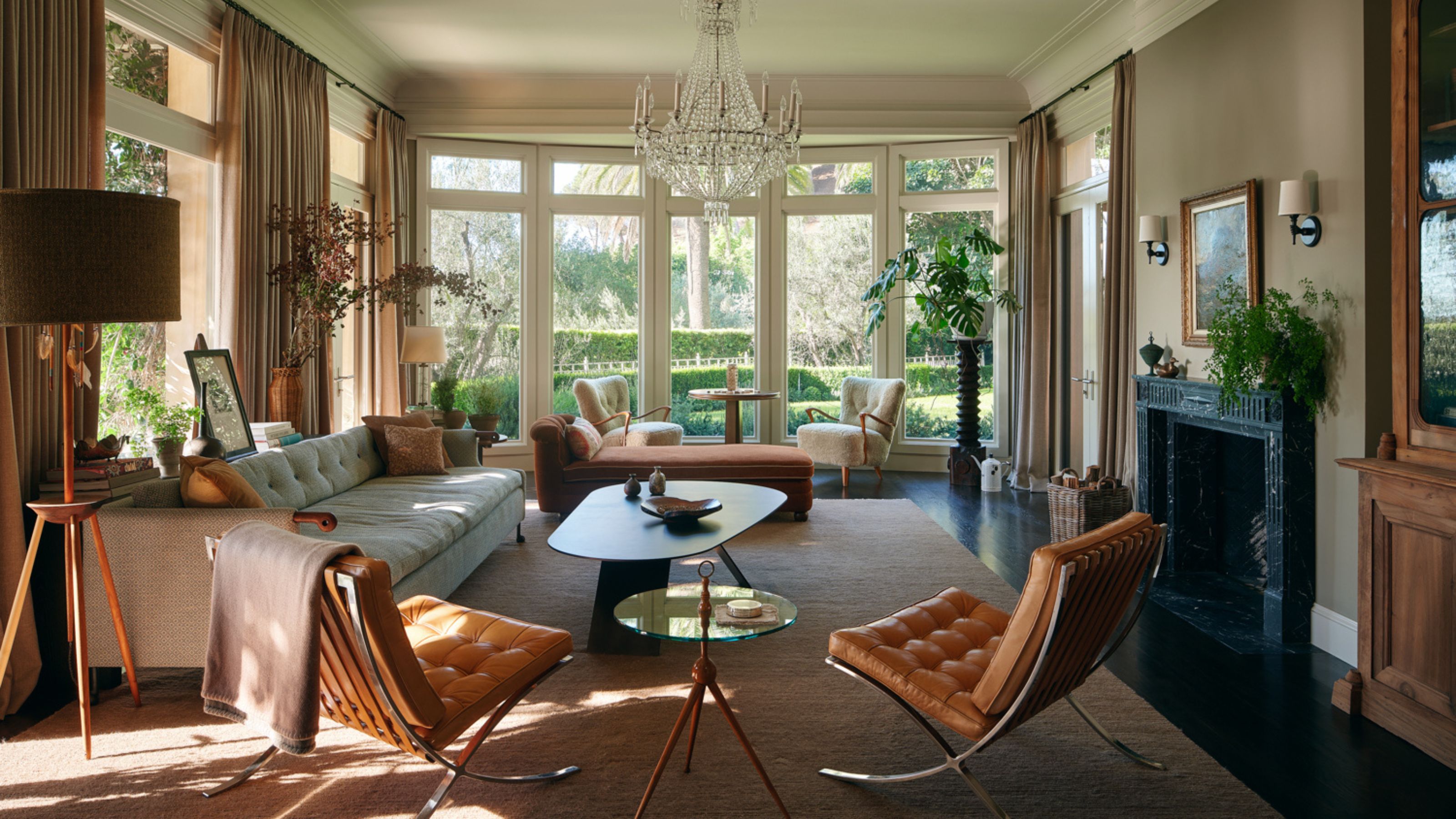 a neutral layered living room with a large bay window overlooking the garden with a glass chandelier, fireplace, leather vintage chairs, a vintage credenza and a dark curved coffee table