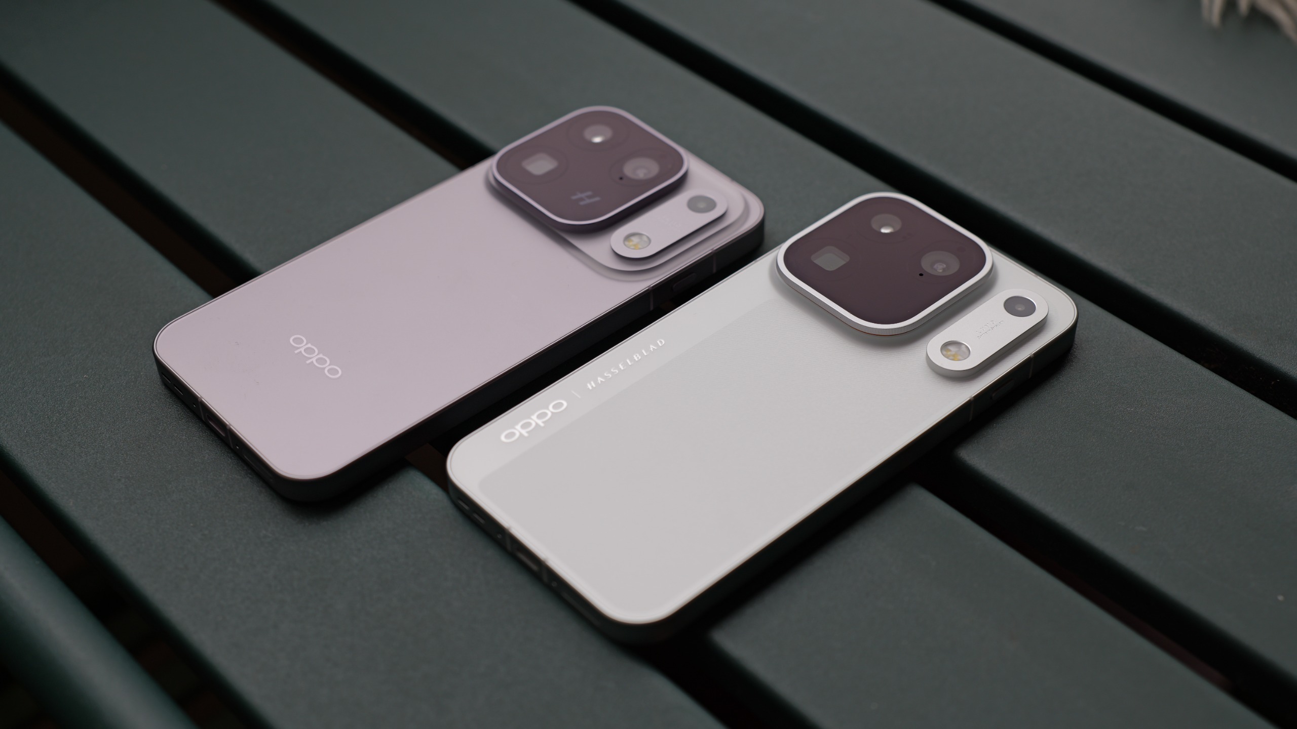 Two Oppo Find X10 series phones lying side by side on a dark green outdoor table, showing white and pale purple finishes.