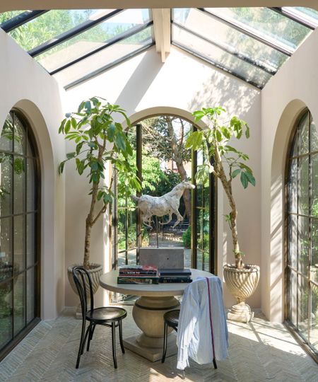 A garden room/orangery with three large glass doors, a circular table in the middle of the room and two large potted trees. The walls are painted in a very pale pink. 