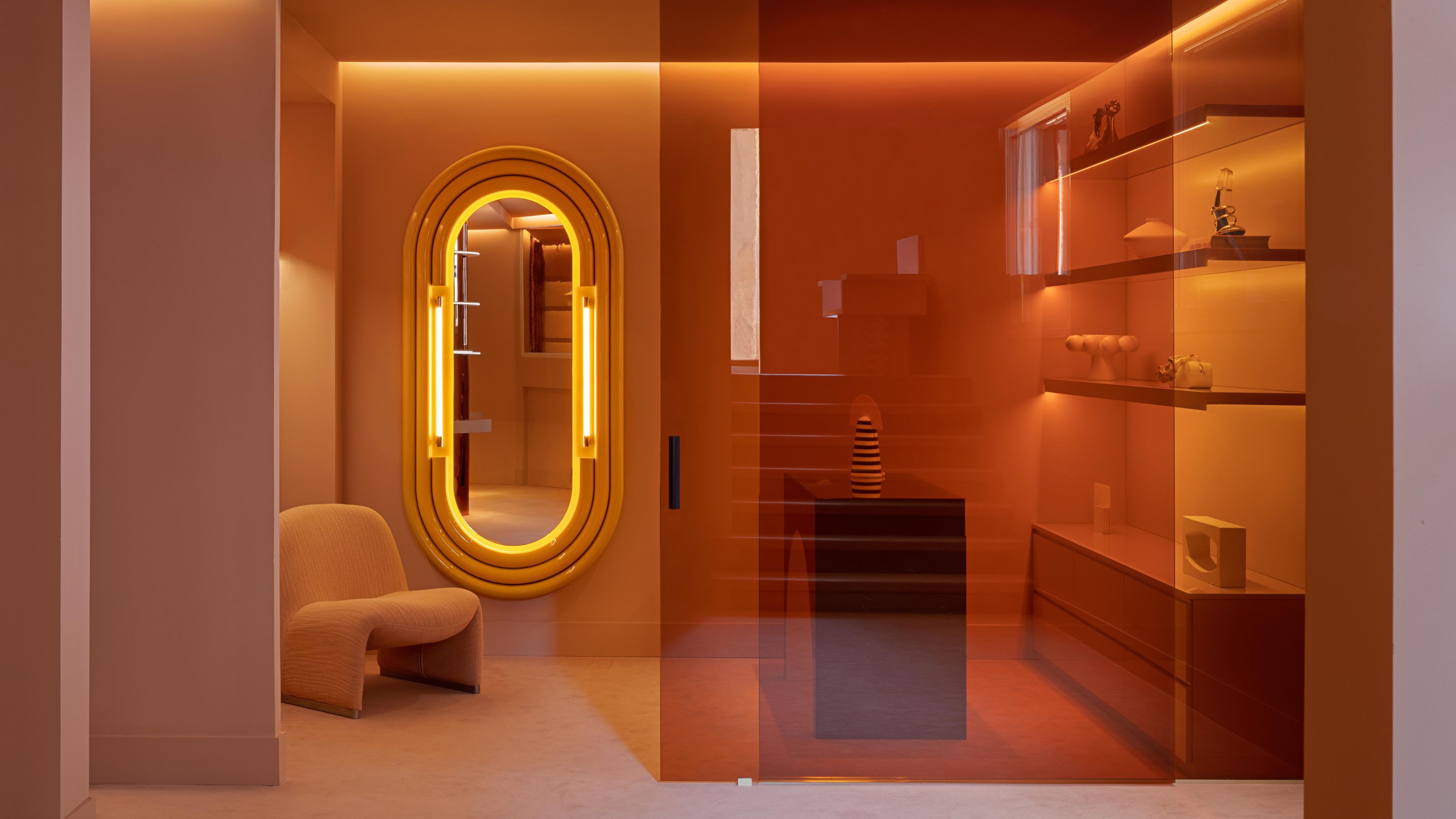 Image of a boutique store room that is color-drenched in a pink peach colorway. There is a futuristic floor-length mirror on the wall, a modern pink accent chair, and a peach glass room divider.