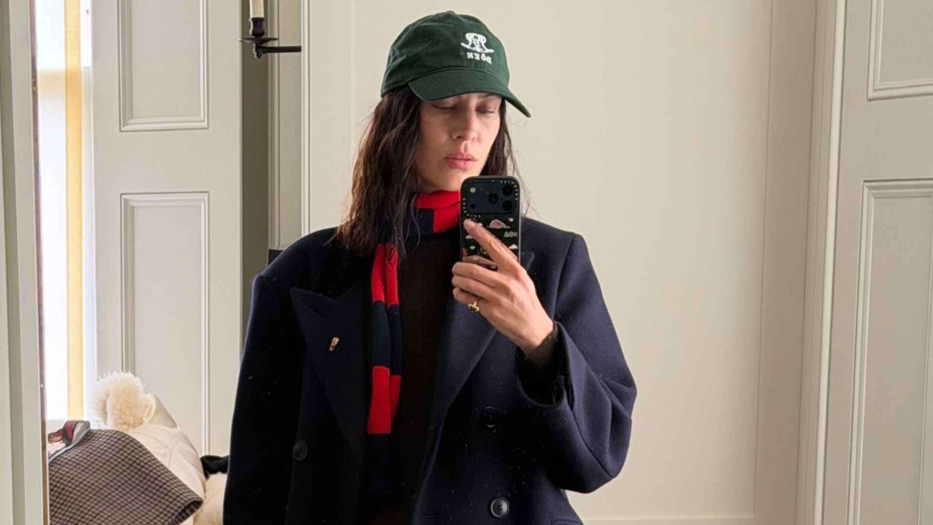 Alexa Chung takes a mirror selfie wearing a navy coat with stove pipe jeans, Tabi loafers and a green cap.
