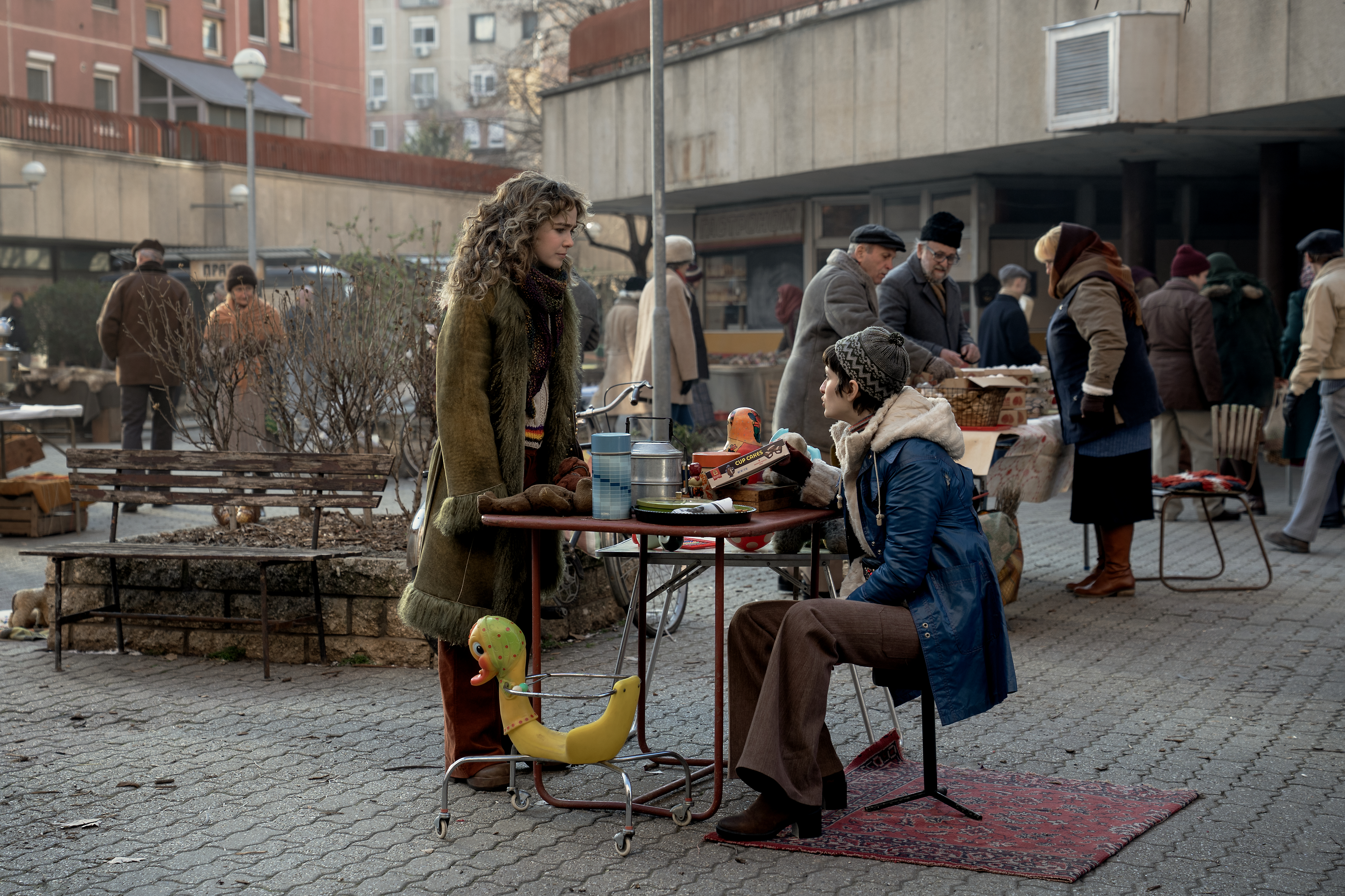 A series of film stills and photographs from a 1970s-inspired set featuring vibrantly decorated interiors with layered furniture and warm lighting, along with characters dressed in vintage clothing whether indoors or at parks, in cards, or at markets outdoors.