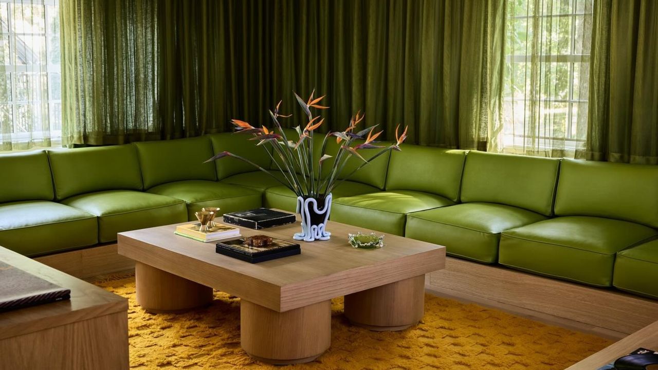 a moody, 70s-inspired living room with a long and low lime green sectional sofa with slightly darker green curtains on the window behind it. There is an orange rug with a wooden coffee table on it.