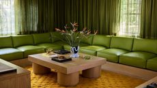 a moody, 70s-inspired living room with a long and low lime green sectional sofa with slightly darker green curtains on the window behind it. There is an orange rug with a wooden coffee table on it.