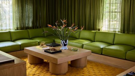 a moody, 70s-inspired living room with a long and low lime green sectional sofa with slightly darker green curtains on the window behind it. There is an orange rug with a wooden coffee table on it.