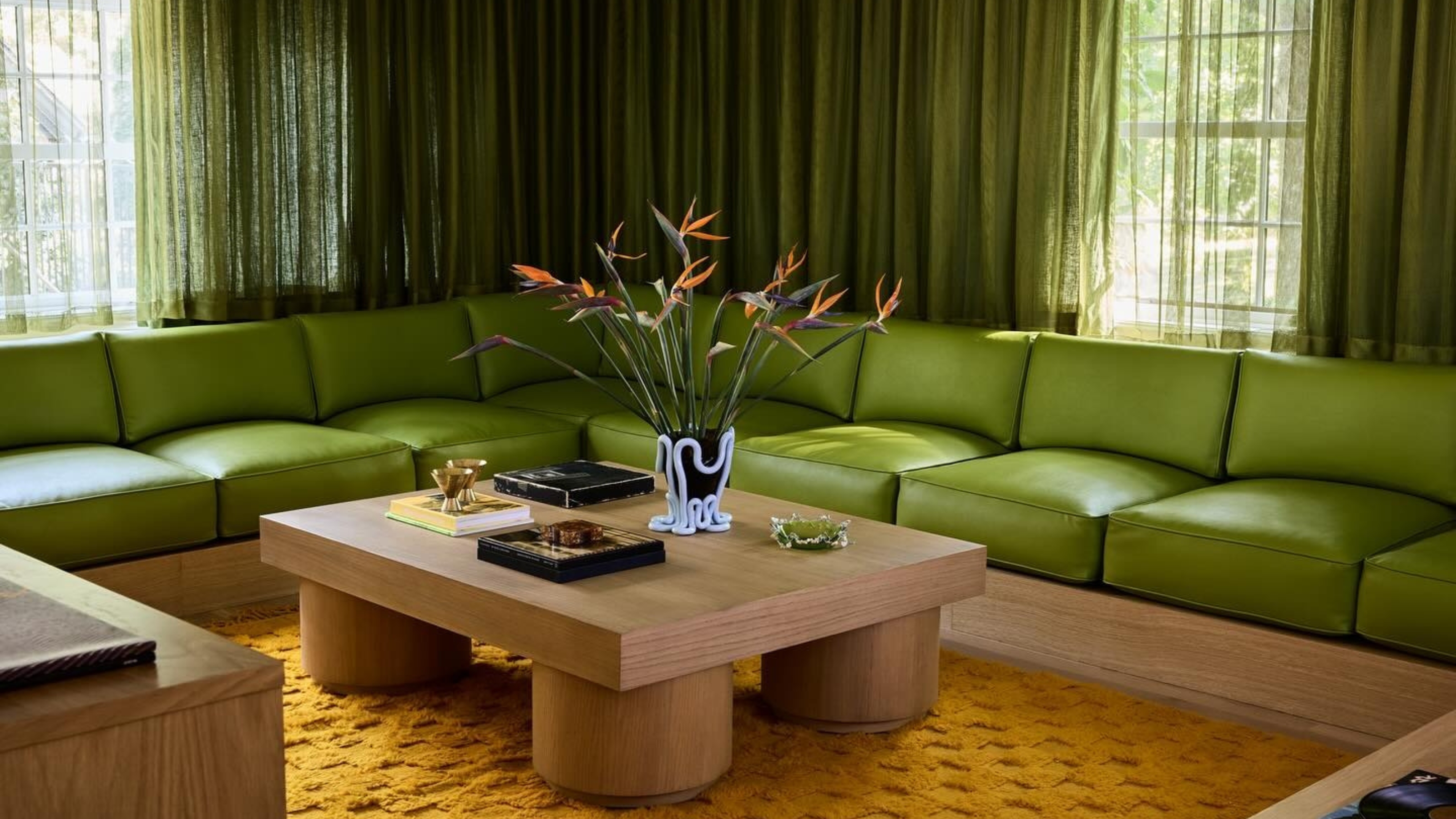 a moody, 70s-inspired living room with a long and low lime green sectional sofa with slightly darker green curtains on the window behind it. There is an orange rug with a wooden coffee table on it.