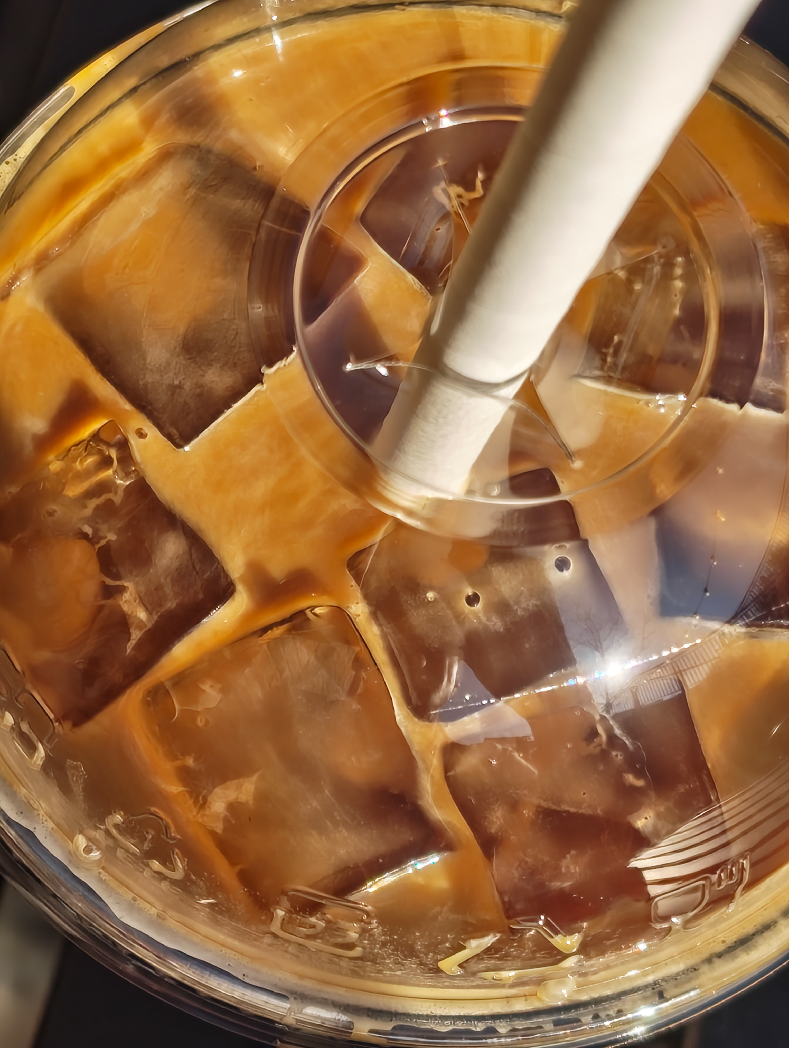 Top-down view of an iced coffee with a straw and melting ice cubes inside a plastic cup, photographed with the Nothing Phone (4a).