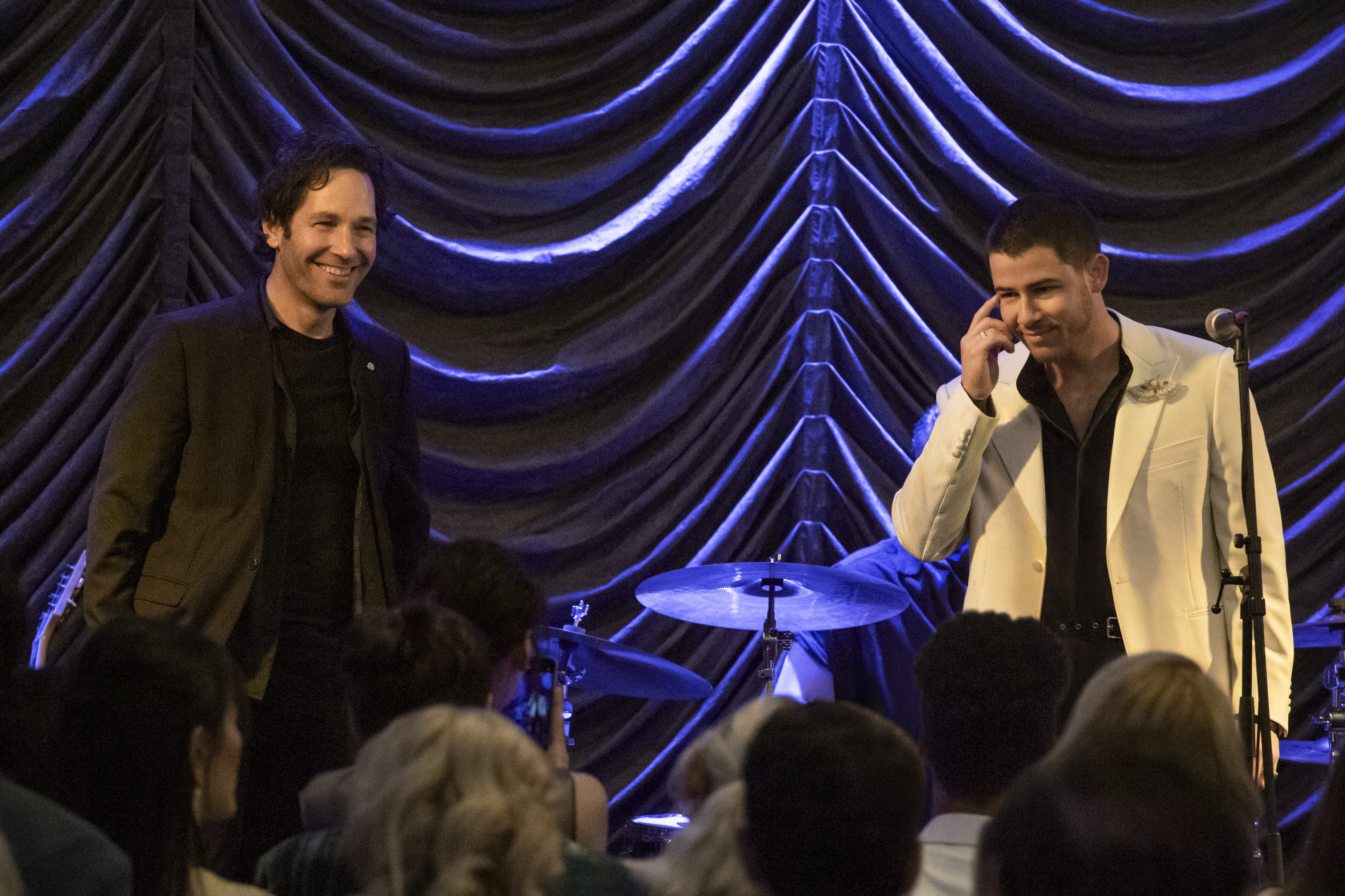 paul rudd and nick jonas smile while on stage at a club in front of a drum set in a still from the movie power ballad
