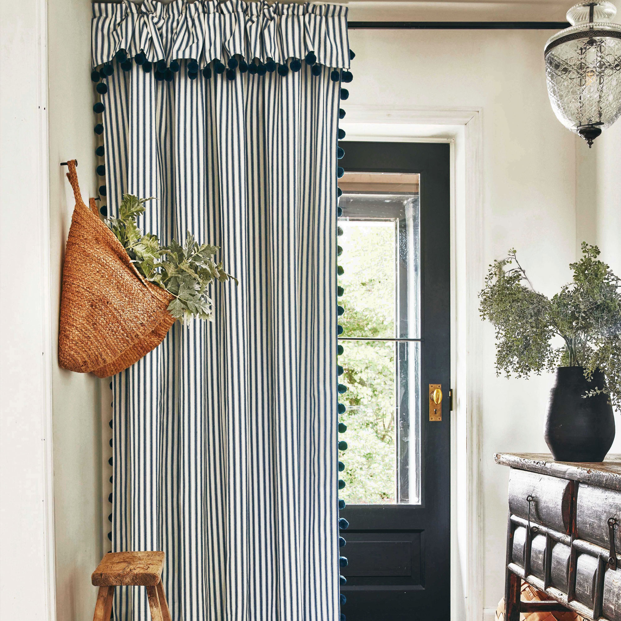 White hallway with blue ticking stripe curtain edged with bobble trim, a basket hung on the wall and a wooden console table