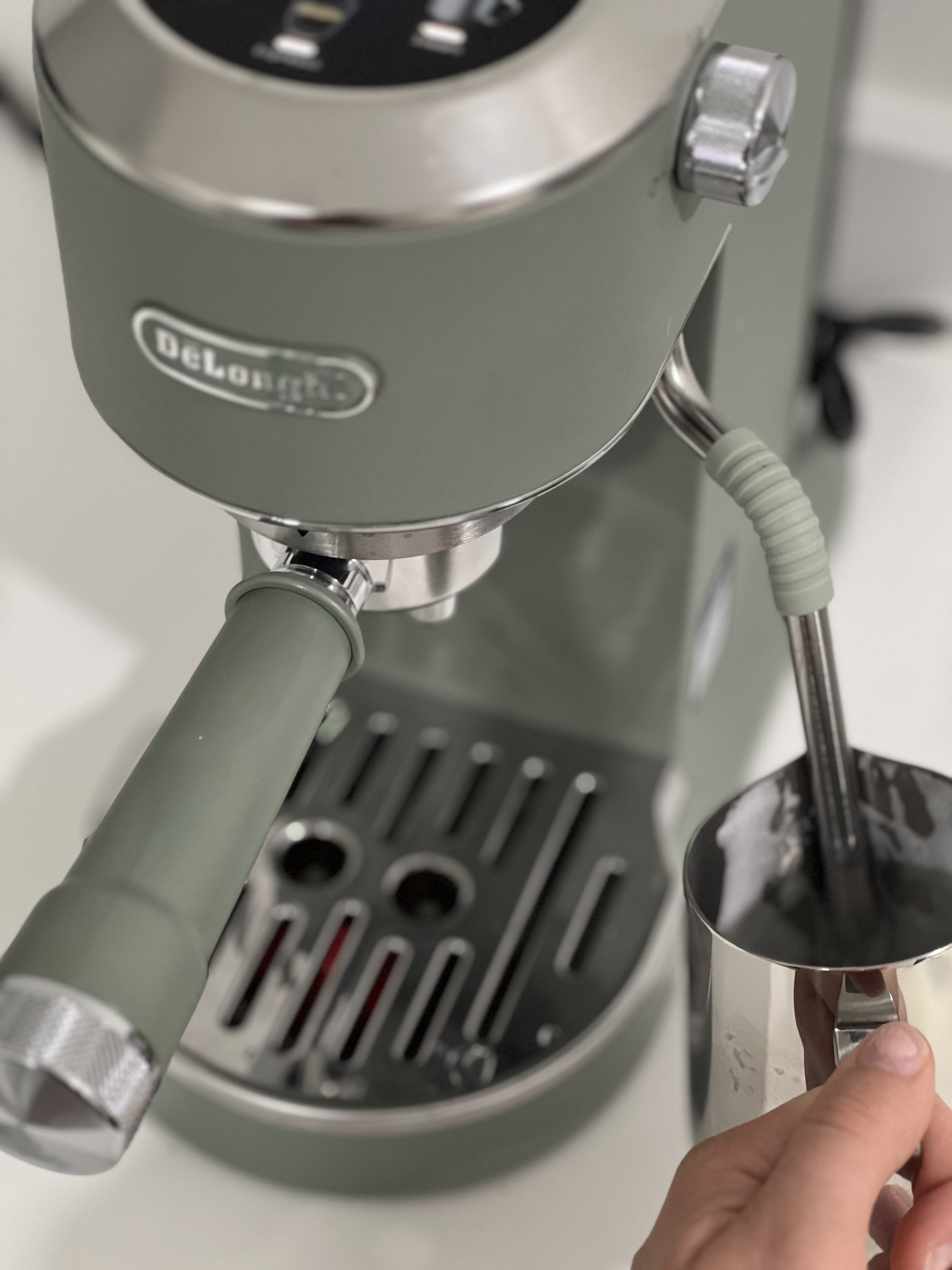 Image of the De'Longhi Dedica Duo espresso machine in sage green with a hand steaming milk in the frother.