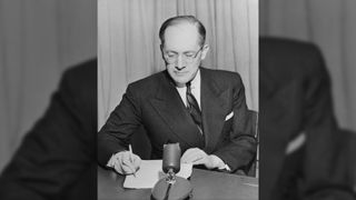 A black and white photograph of international lawyer Raphael Lemkin as he signs important documents. He is a balding man with a combover, and is wearing round glasses and a pin-striped suit and square-patterned tie.