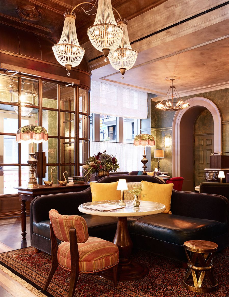 A warmly lit, retro-inspired hotel lobby boasts plush fabrics and rugs, styled with transitional chandeliers, Art Deco table lamps, and whimsical sculptures.