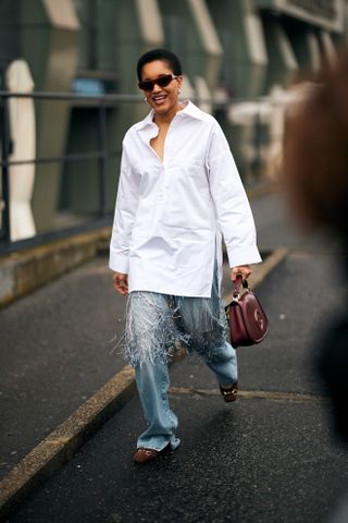 Tamu McPherson at Milan Fashion Week Fall 2025 street style in wide-leg blue jeans, a white blouse, red bag, and brown suede loafers.