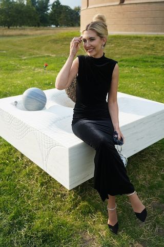 Ashley sits on top of her art piece wearing a long black dress.