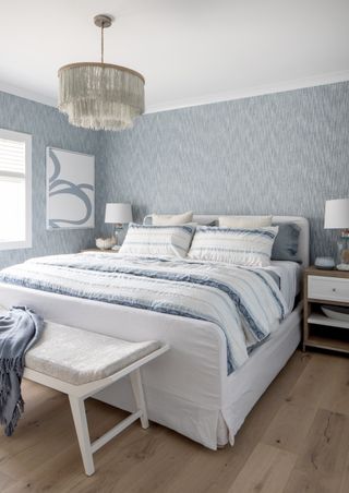 bedroom with pale blue textured wallpaper, blue and white bedding/blanket, white bed, bench, side table, artwork, table lamps and pendant, wooden floor
