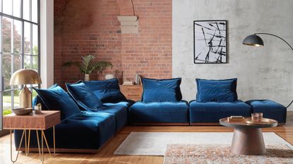 A modern industrial living room with blue velvet L-shaped sofa, exposed brick wall and laminate flooring