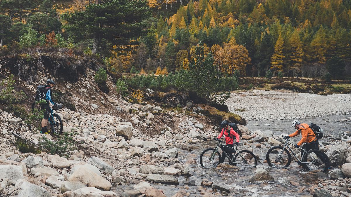 Three bikepackers pushing their bikes across a river in Scotland