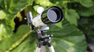 A lens-on view of a telescope standing on a tripod in front of large green leaves.