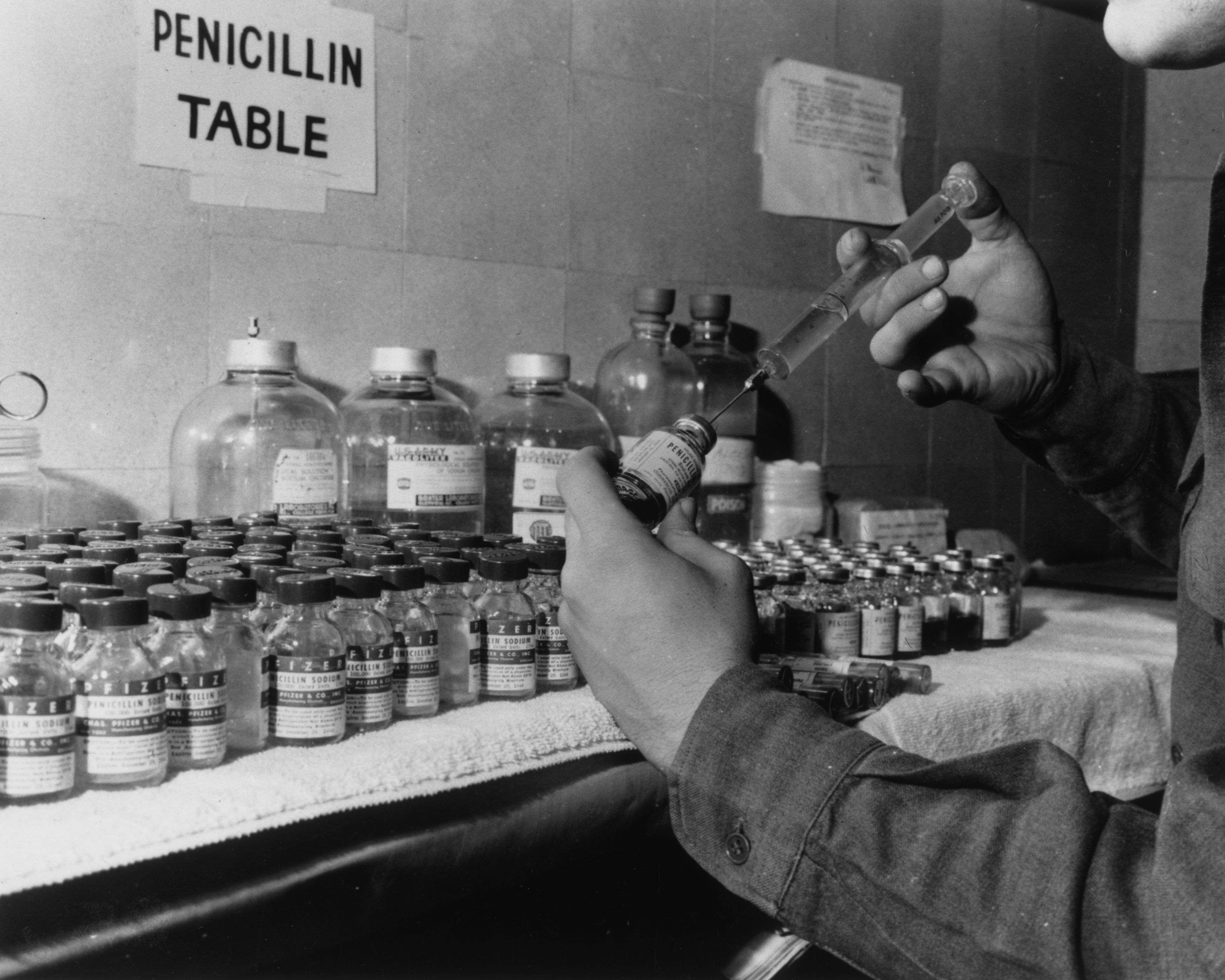 A view of a man filling a syringe with penicillin in front of a counter filled with penicillin vials, 1940s.