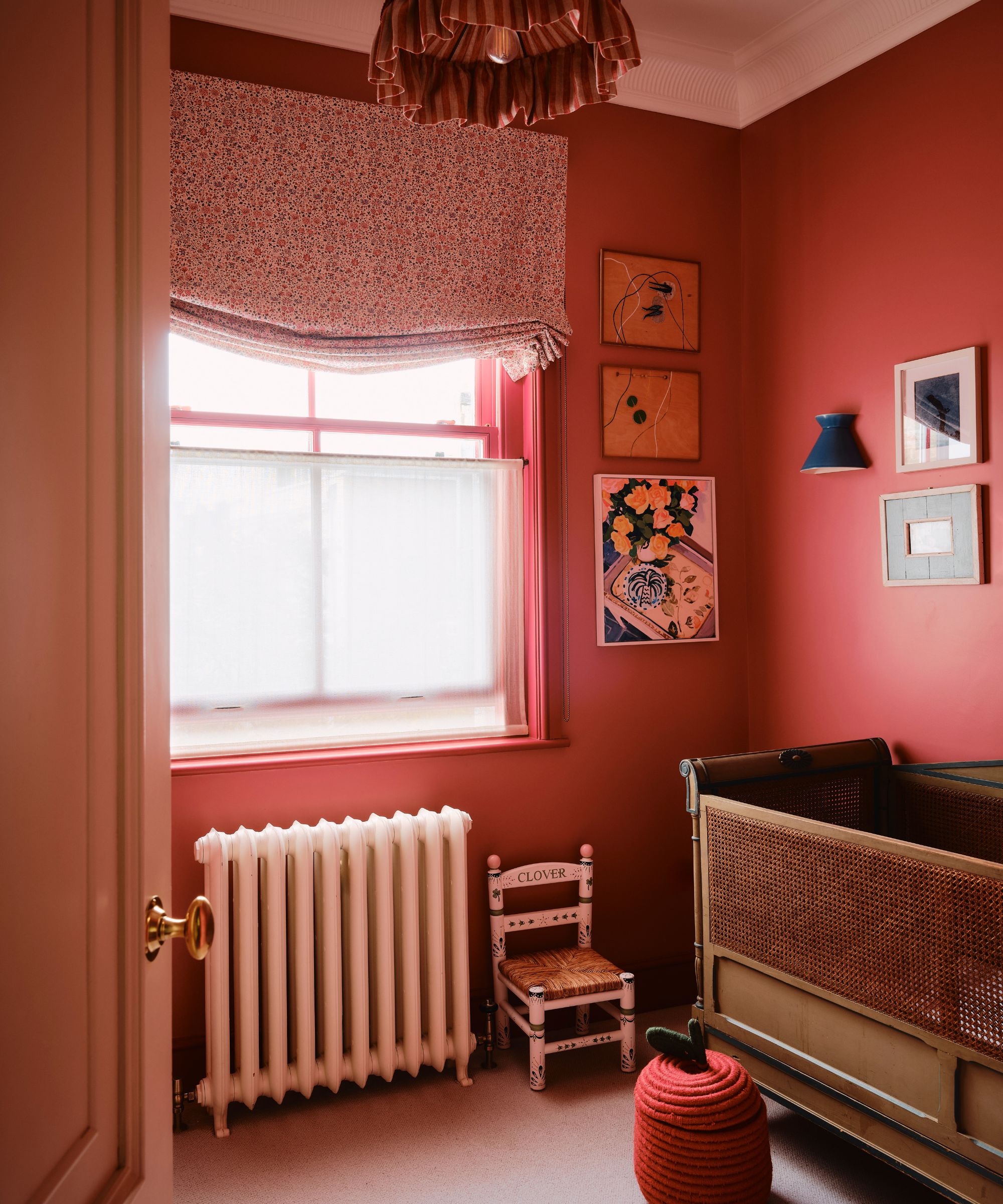 small dark pink nursery bedroom with a large antique cot, roman blind and cafe curtain at the window, a small painted chair and eclectic wall art
