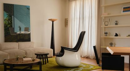 A modern apartment with a cream modular sofa, acid green tufted rug, a sculptural chair in lacquered ceramic and black leather, a standing lamp, minimal shelving, and white curtains.
