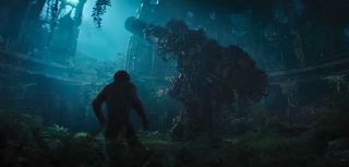 an ape stands next to a big, weed-covered, floor-mounted gun inside a decrepit building overgrown with plant life.