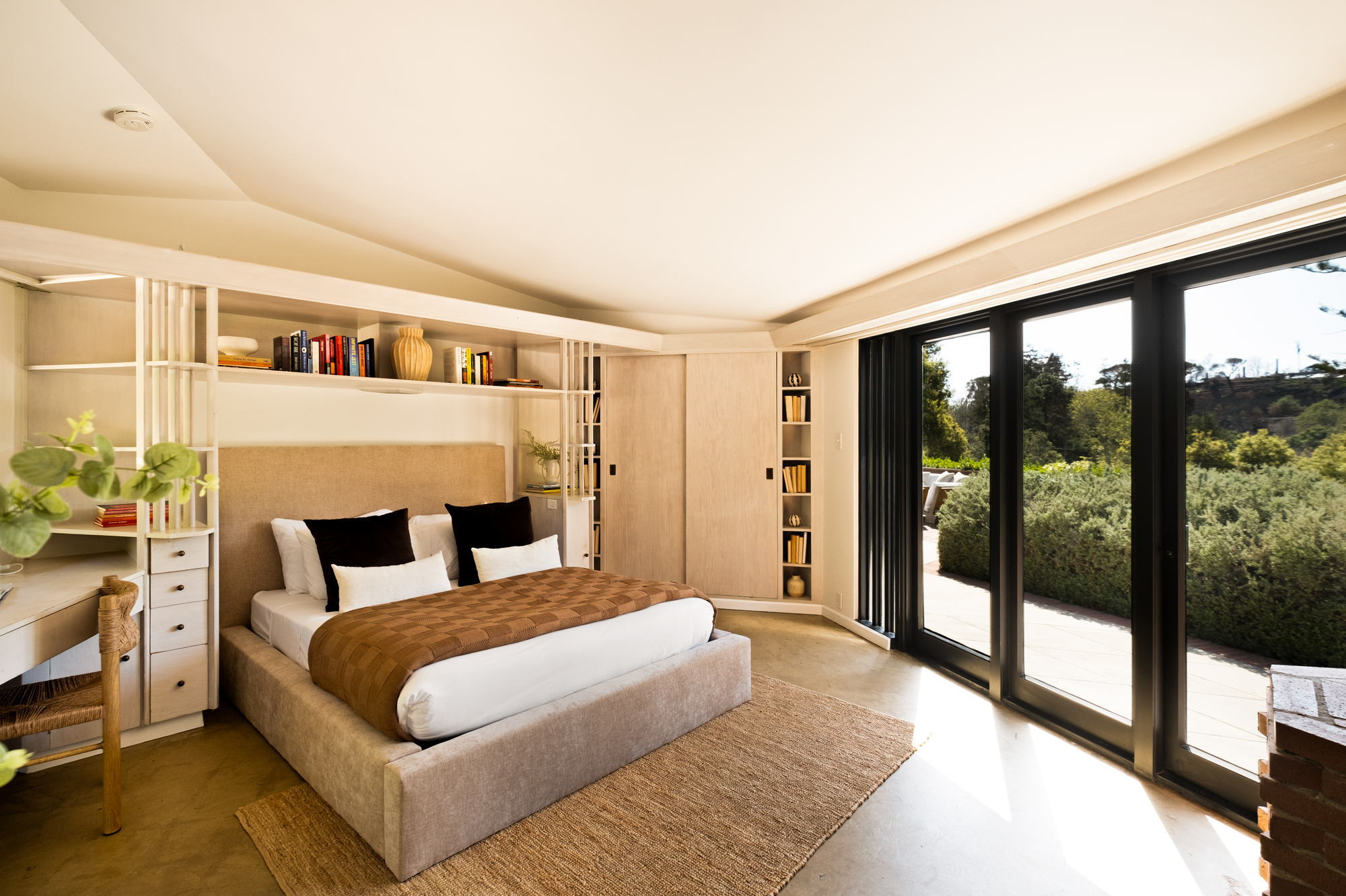 Image of an angular bedroom with a tan upholstered box bed. There is built-in shelving around the bed and a desk to the left. The wall to the right is made of glass doors that lead to the outdoors.