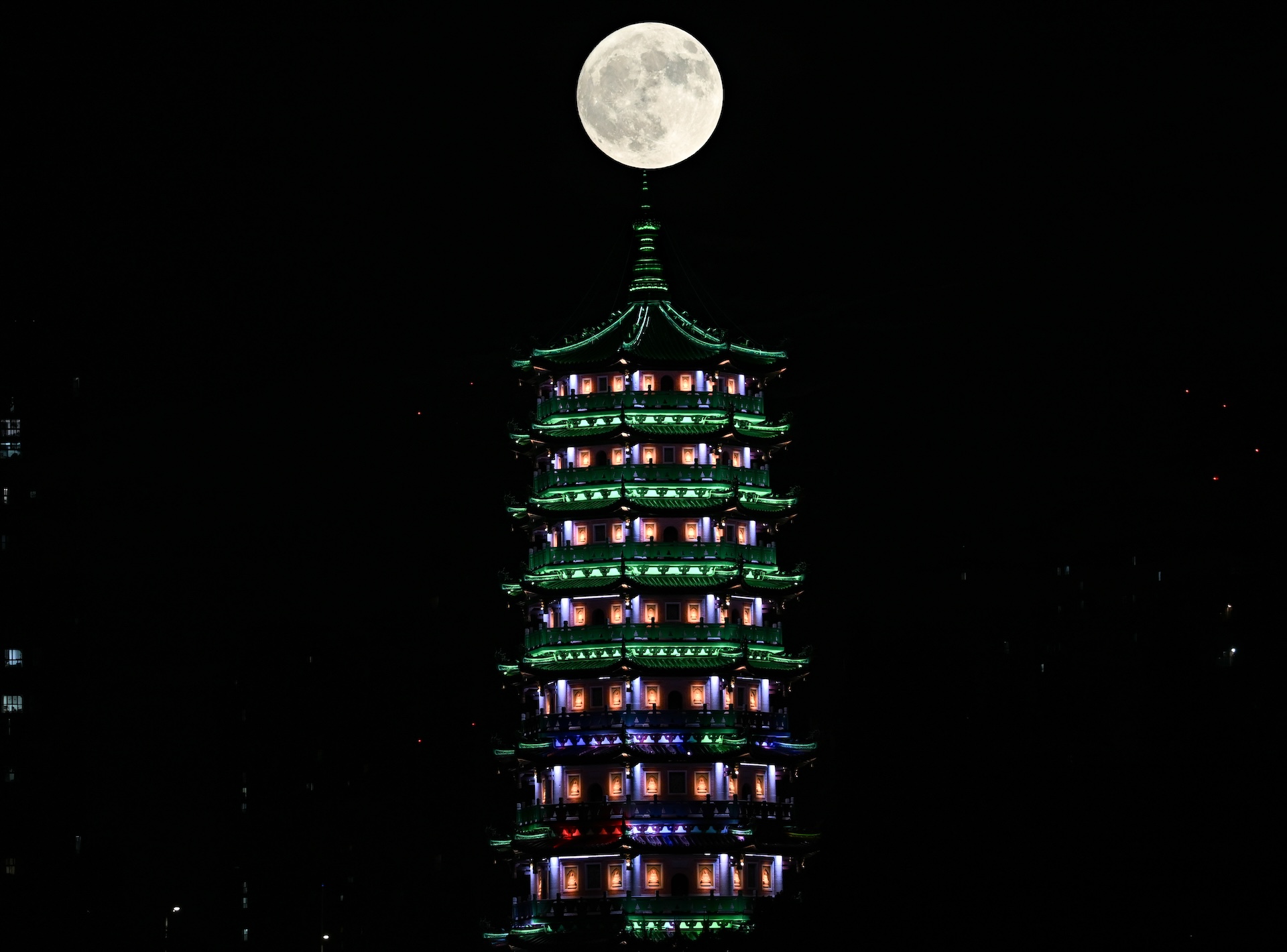 Uma lua branca se alinha perfeitamente acima de um templo alto iluminado por luzes coloridas