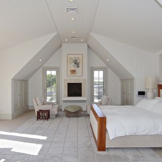 A white bedroom with vaulted ceiling containing a white bed, armchairs and rug.