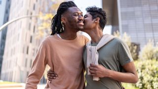 Man kisses another man on his cheek while he has his arm wrapped around his waist. The man who is being kissed is smiling.