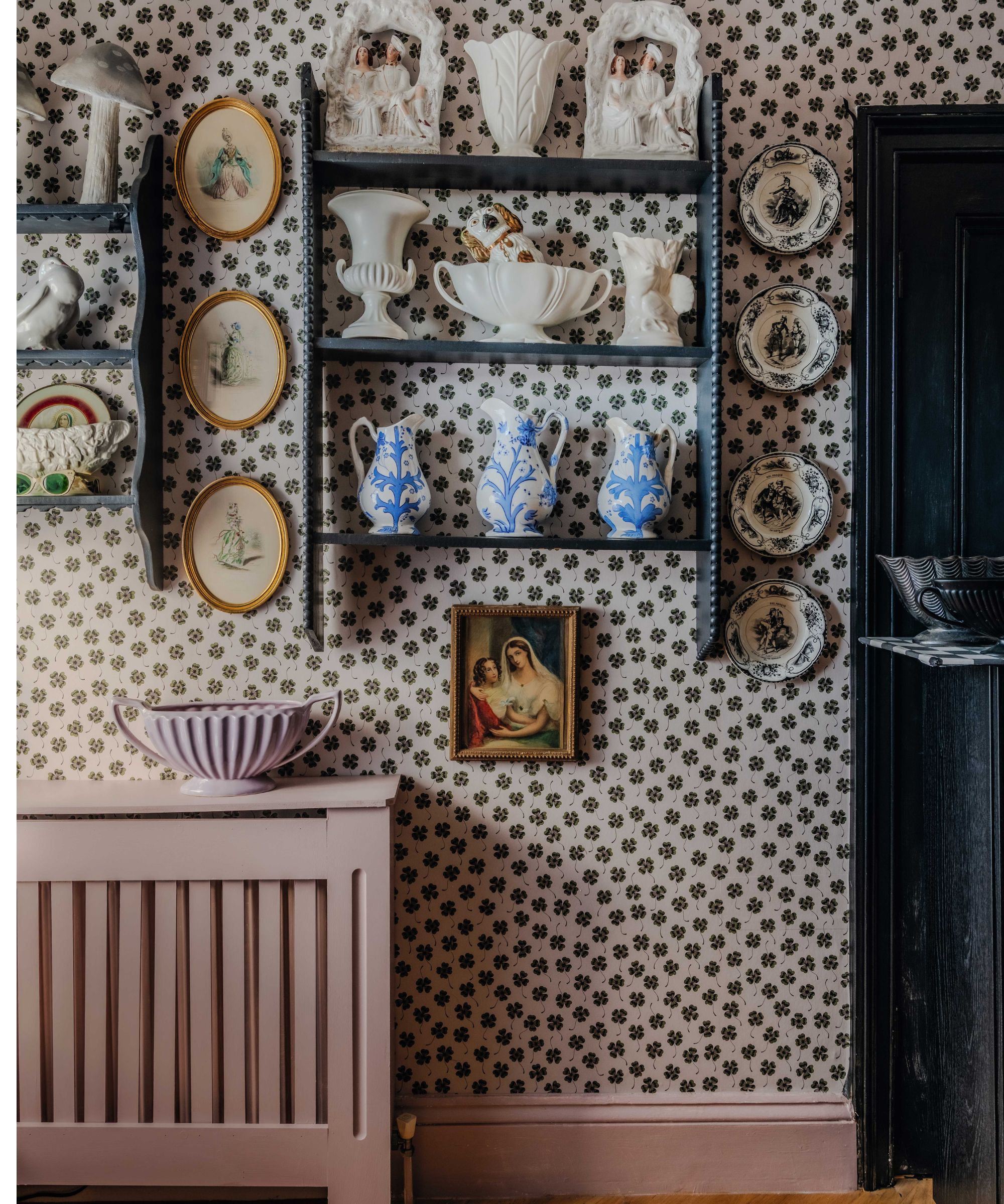a floral wallpapered sitting room with dusty pink painted skirting and a black painted door with wall shelves filled with vintage collections and wall plates