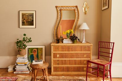A textured bamboo dresser topped with a unique, wavy-framed rattan mirror, yellow daffodils in a glass vase, and a pink marble lamp against a beige wall.