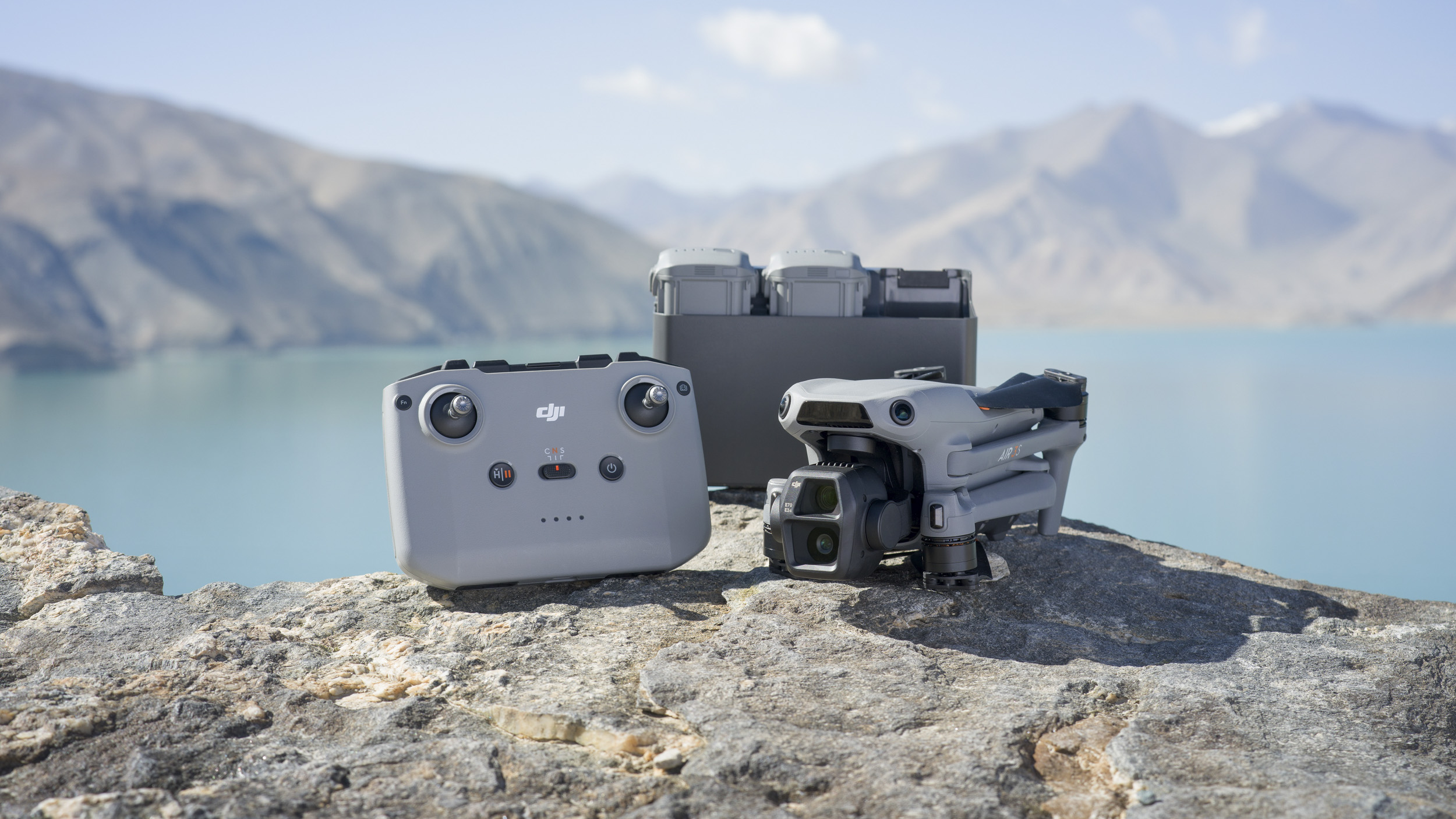 DJI Air 3S drone alongside remote controller and battery charging hub, on a rock with glacial lack and mountain backdrop