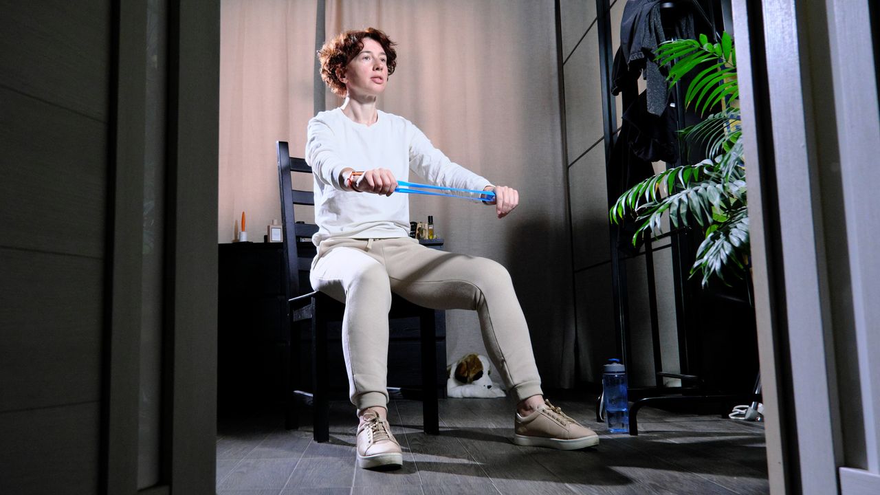 Woman sits in a chair in a domestic setting, holding a small blue resistance band above her knees. She is wearing a white long-sleeved top, beige pants and beige trainers.