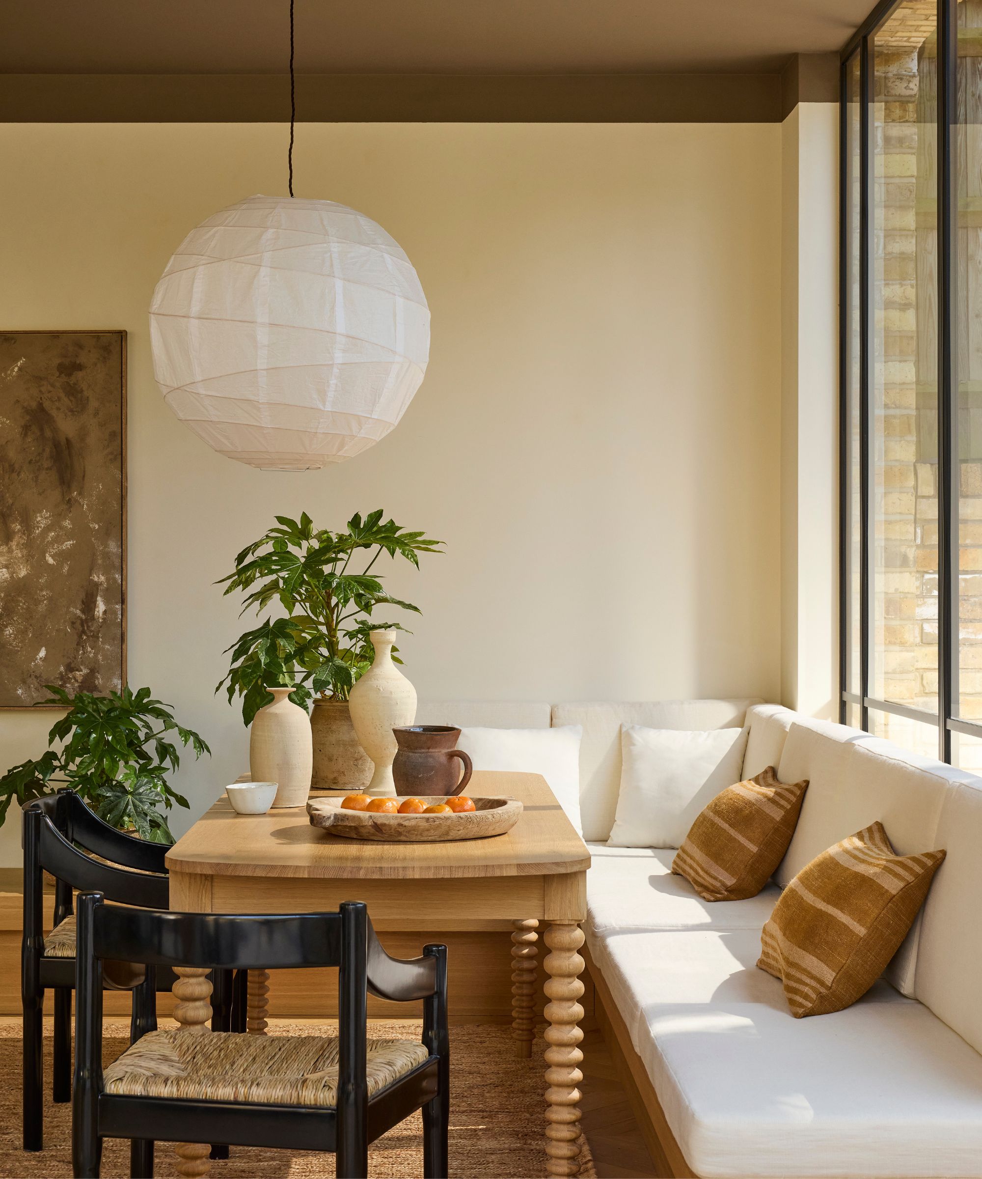 Neutral breakfast nook with cream walls, a cream built-in banquette, a chocolate-brown ceiling, a white lantern pendant light, a wooden bobbin-leg table, and black wicker dining chairs.