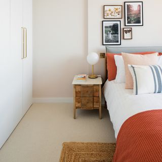 A contemporary guest room with wooden bedside table, white wardrobes with brass handles, framed wall art, jute rug and bed with rust orange throw and pillow decor