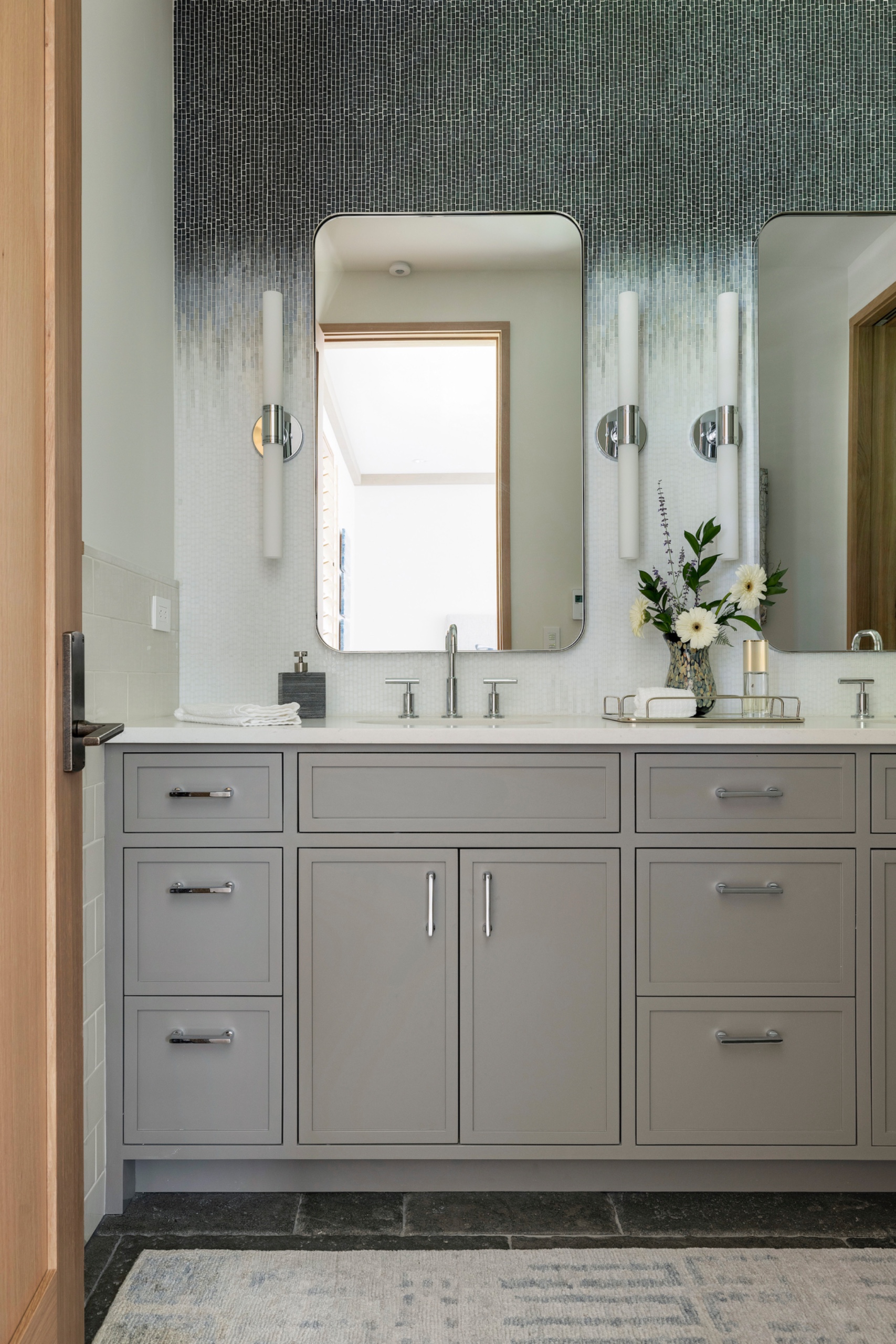 Bathroom with a gray double vanity, tall mirrors and chrome wall lights against a mosaic wall fading from charcoal to white.