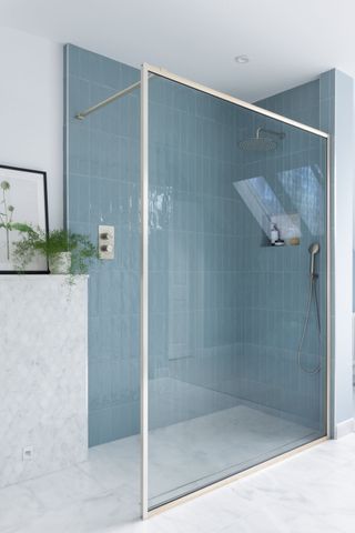 A walk-in shower featuring glossy blue vertical tiles paired with a sleek, gold-framed glass panel. The shower area is complemented by a marble-tiled partition, a built-in niche for toiletries, and a rainfall showerhead. Natural light pours in through a skylight, enhancing the airy and minimalist design.