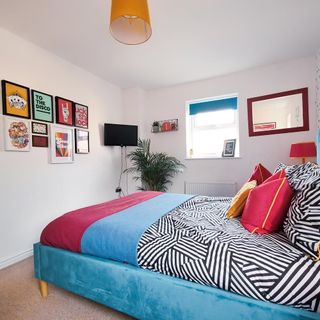 White bedroom with blue bed, striped duvet, pink cushions, wall pictures and orange pendant light