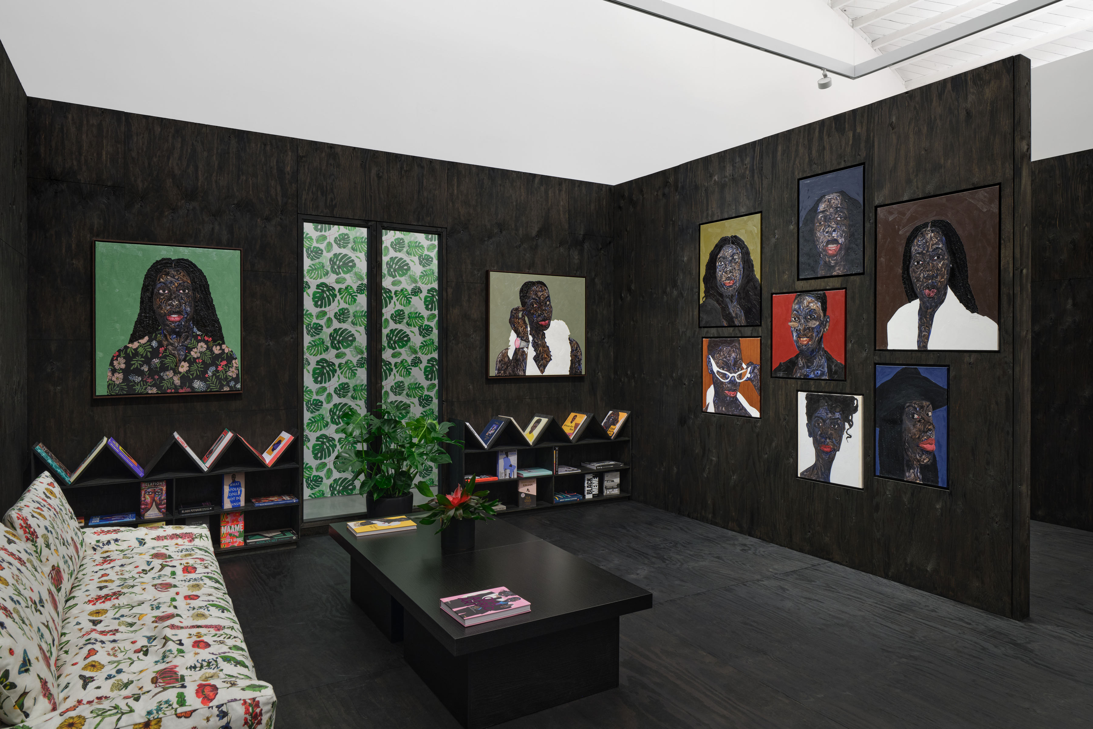 View of an art gallery installation with dark wood walls and multiple expressive portrait paintings. In the foreground, a large black metal structure with vertical bars frames additional paintings inside, while more portraits hang on the walls in the background.