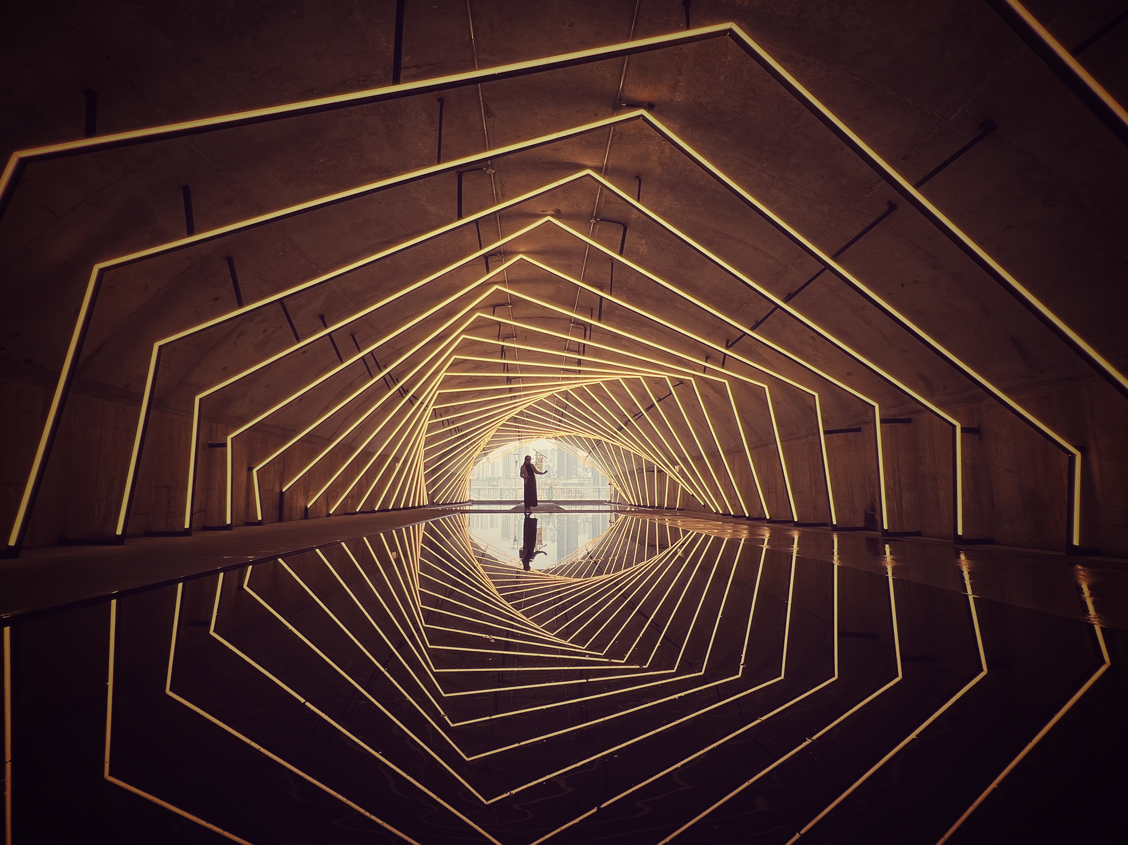 A person stands in a tunnel lined with glowing geometric patterns. The reflection on a glossy floor creates an infinite loop effect