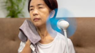 A middle aged woman with cropped black hair sits on a couch and wipes her face with a small towel, as if overheated. A small electrical fan is in the foreground and is pointed at the woman.