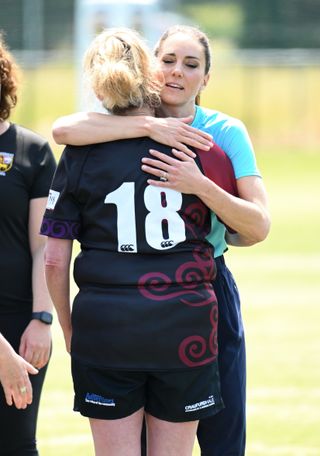 Kate Middleton standing in a grass rugby field hugging Sarah Renton who is wearing a black jersey and shorts