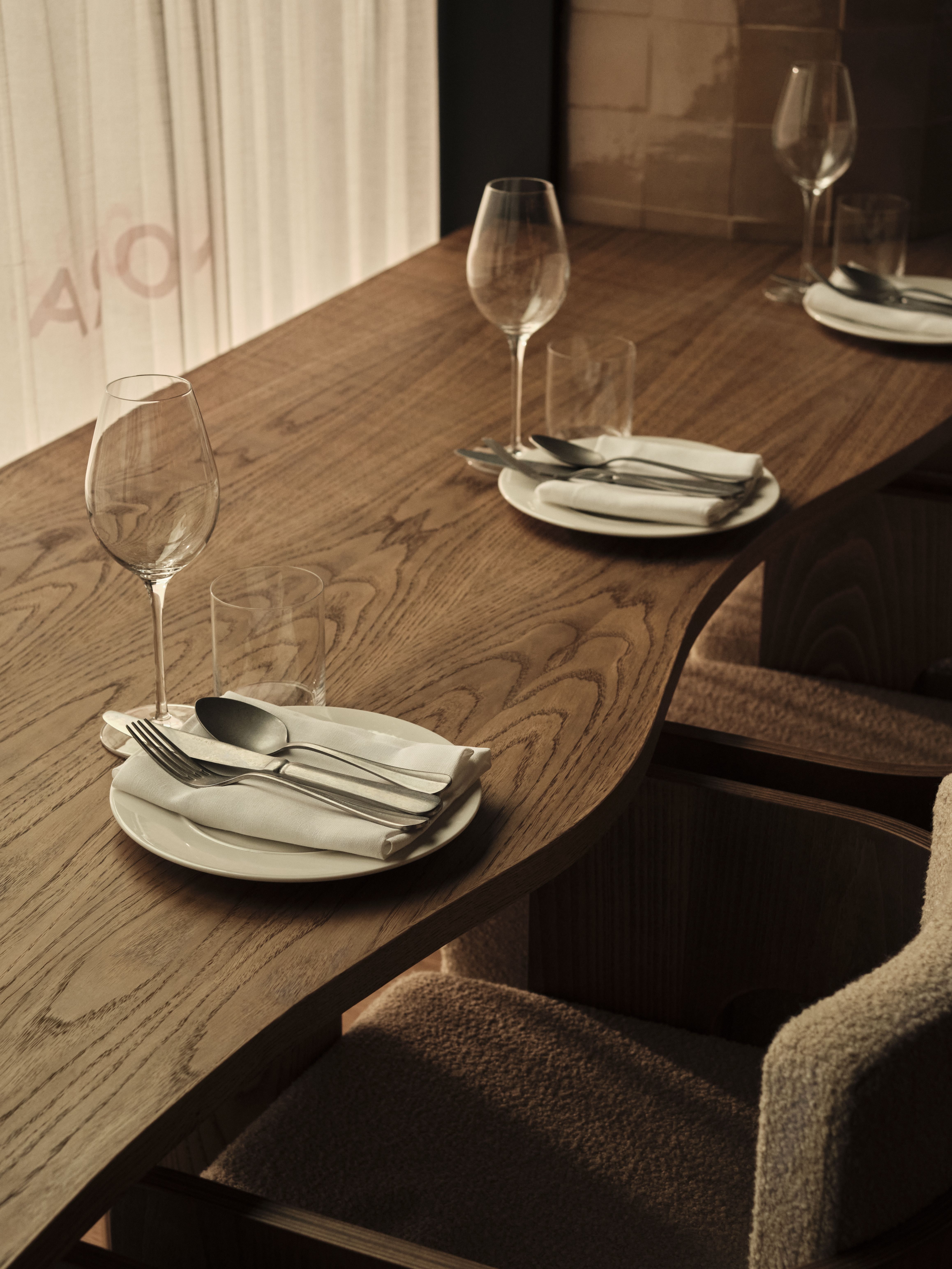 A restaurant's wooden counter with wavy silhouettes is paired with upholstered beige shearling and wood stools and classic tableware.