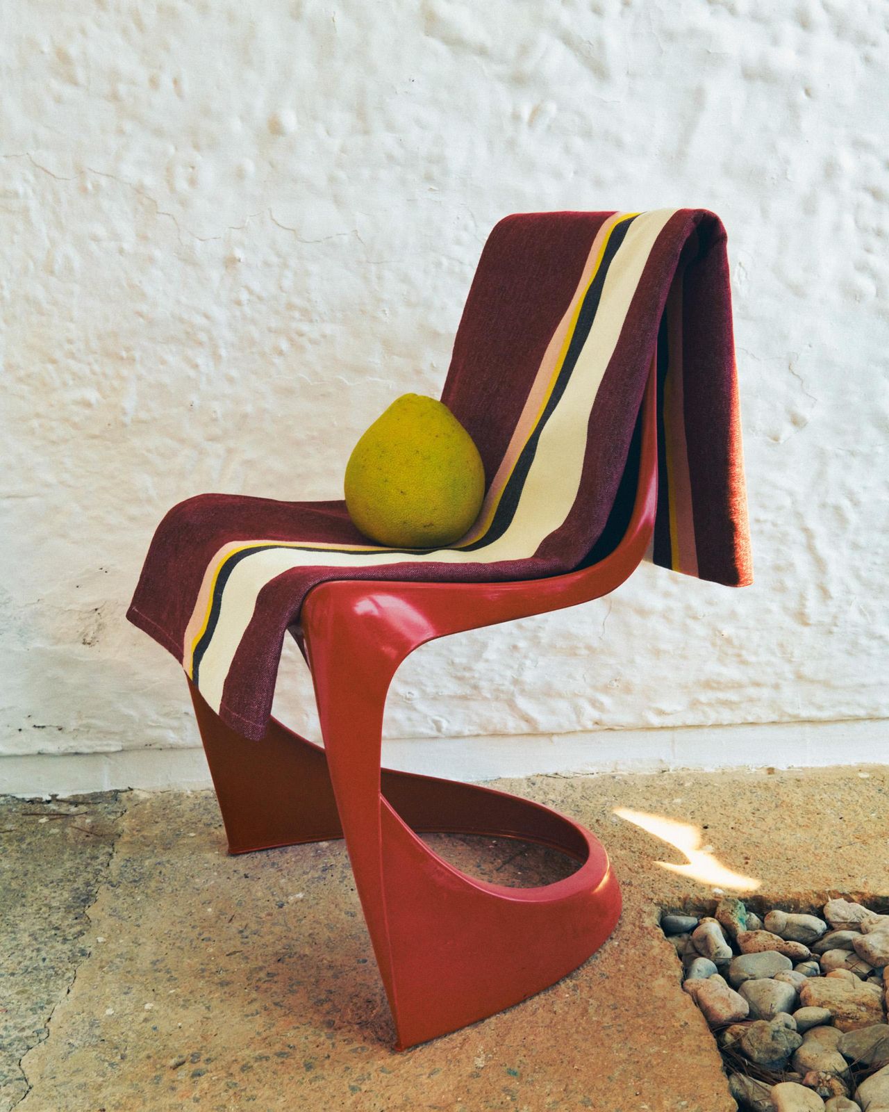Image of a red chair with a burgundy, yellow, black, and white striped towel draped over it. There is a mango on the chair as well.