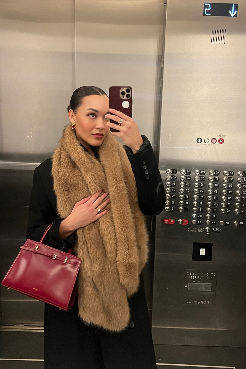 West Village winter trends are shown in a close street-style photo of a woman standing in an elevator taking a selfie in her winter outfit. She's wearing gold earrings with a long black coat, a brown fur scarf, and a red structured handbag