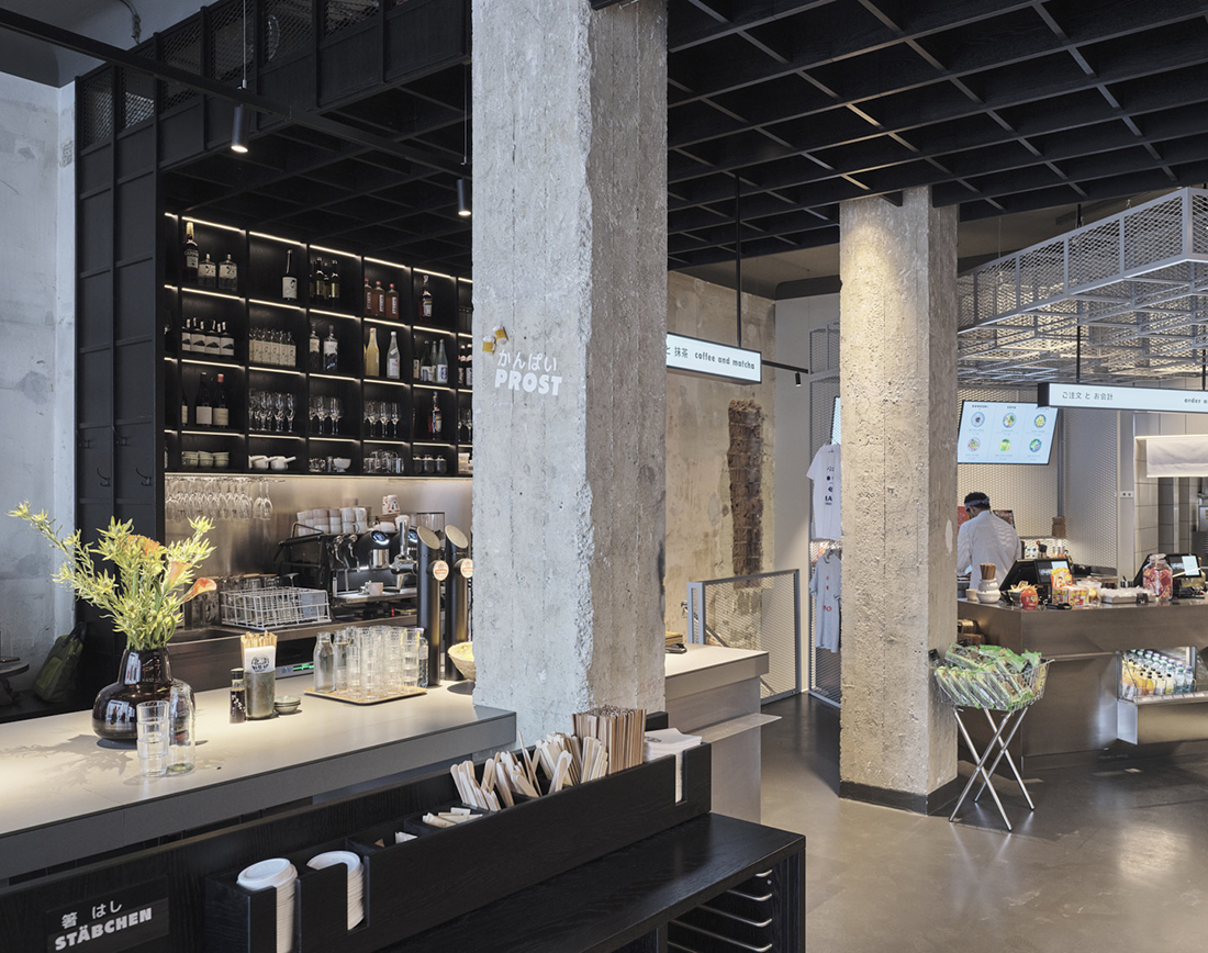 A grocery store with brutalist-like interiors, including exposed cement pillars, black wooden or iron furniture, and bright lighting.