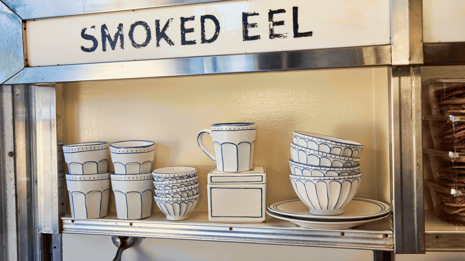 A rotating gif showcasing the warm, old-style interiors of a deli, with displayed bagels, pastries, and bread, white and blue ceramics, and white-jacketed staff smiling.